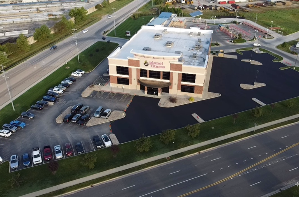 Aerial view of a Planet Fitness gym building with a commercial asphalt parking lot, several parked cars, and nearby roads with light traffic; green grassy areas and trees surround the property.