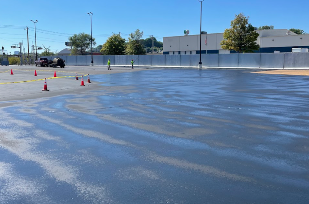 A freshly paved parking lot in St. Louis, MO with wet asphalt is cordoned off by orange cones and caution tape. Several workers in safety vests from local Asphalt Services are visible under a clear blue sky.