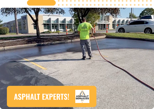 A worker in a neon green shirt uses a hose to spray asphalt in a parking lot near a modern building in St. Louis, MO. Text reads ASPHALT EXPERTS! with the Asphalt Services logo in the bottom right corner.